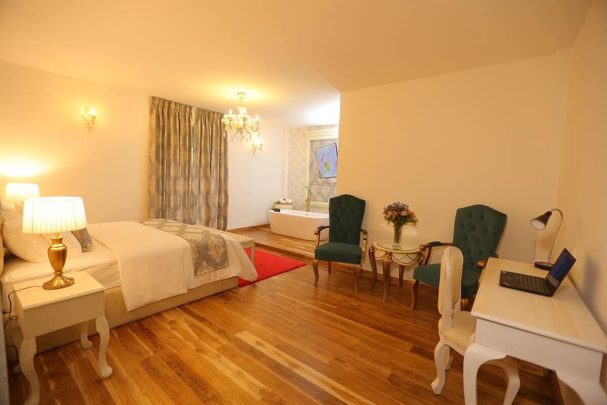 A hotel room with a bed, a desk with a laptop, two chairs, a small table with a vase, and a window with curtains.