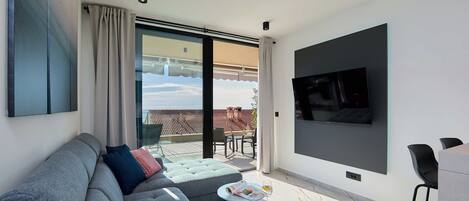 A modern living room with a grey sofa, a small round table with a magazine and a glass of wine, and a flat-screen TV mounted on the wall.