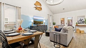 A modern living room with a grey sofa, a wooden dining table set for four, and a flat-screen TV mounted on the wall.