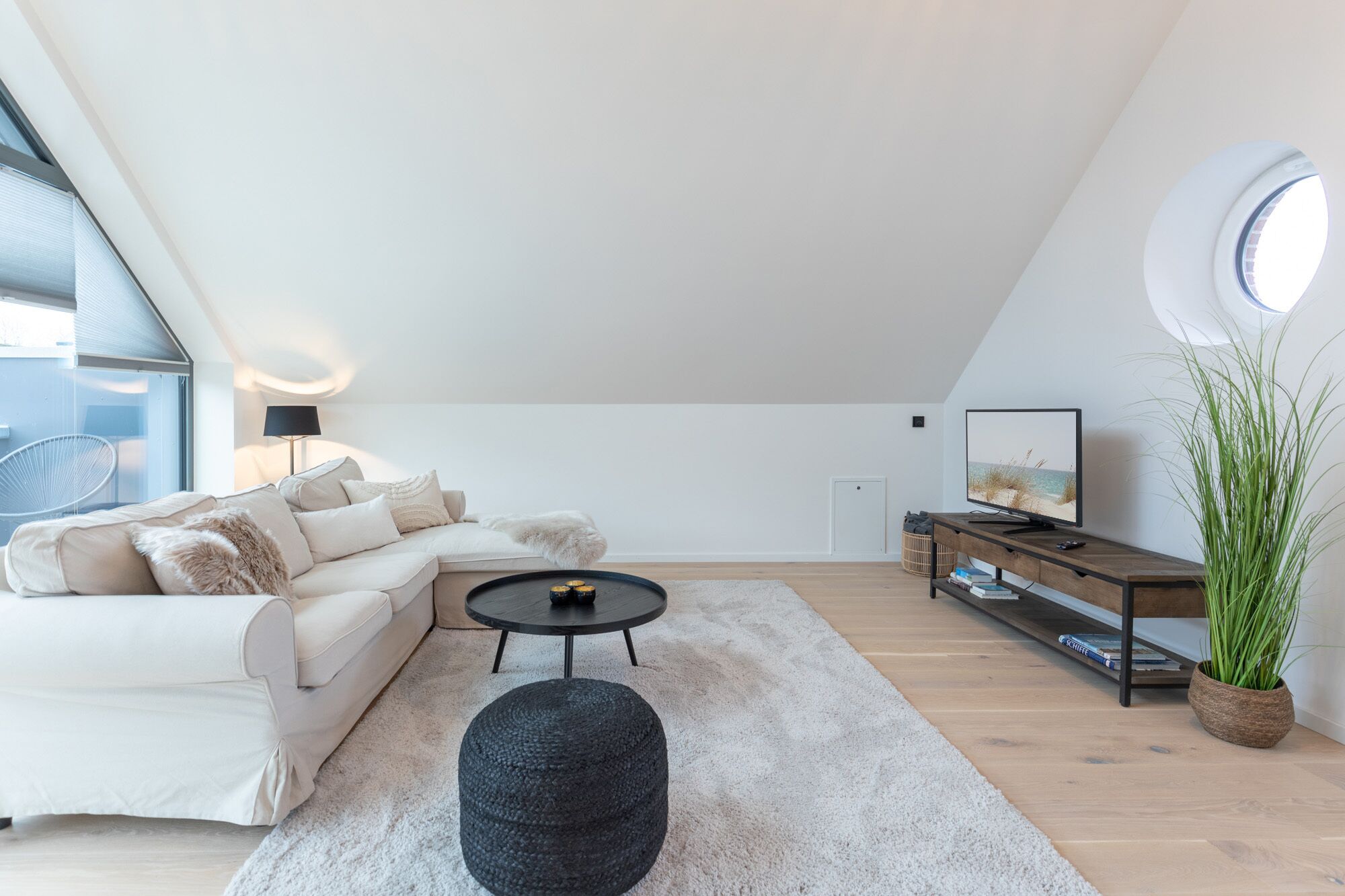 A modern loft bedroom with a large skylight, a sofa, a round ottoman, a TV on a wooden console, and a potted plant.