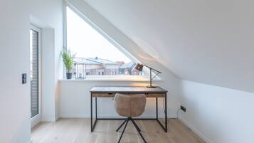 A modern loft bedroom with a sloped ceiling, a wooden desk, a chair, and a large window overlooking a residential area.