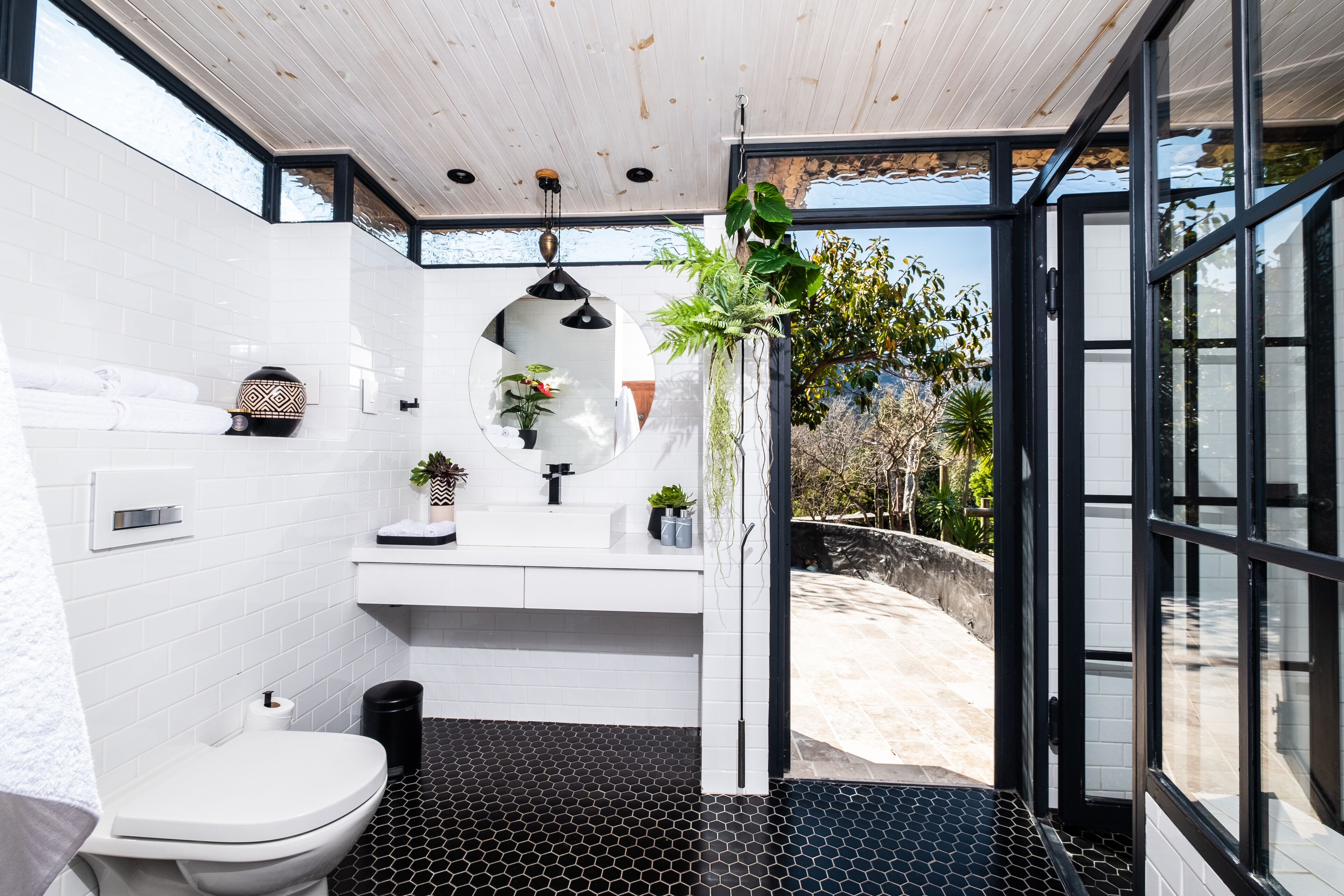 Un baño moderno con inodoro blanco, baldosas hexagonales negras en el suelo, un lavabo blanco con espejo y un cubo de basura negro.