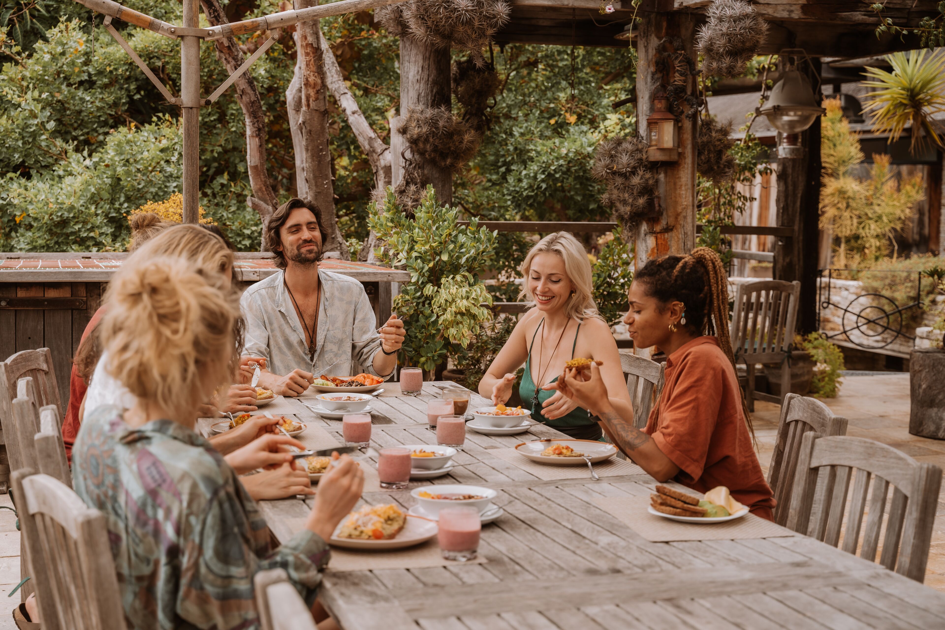 A group of people dining at an outdoor wooden table with various dishes and drinks.