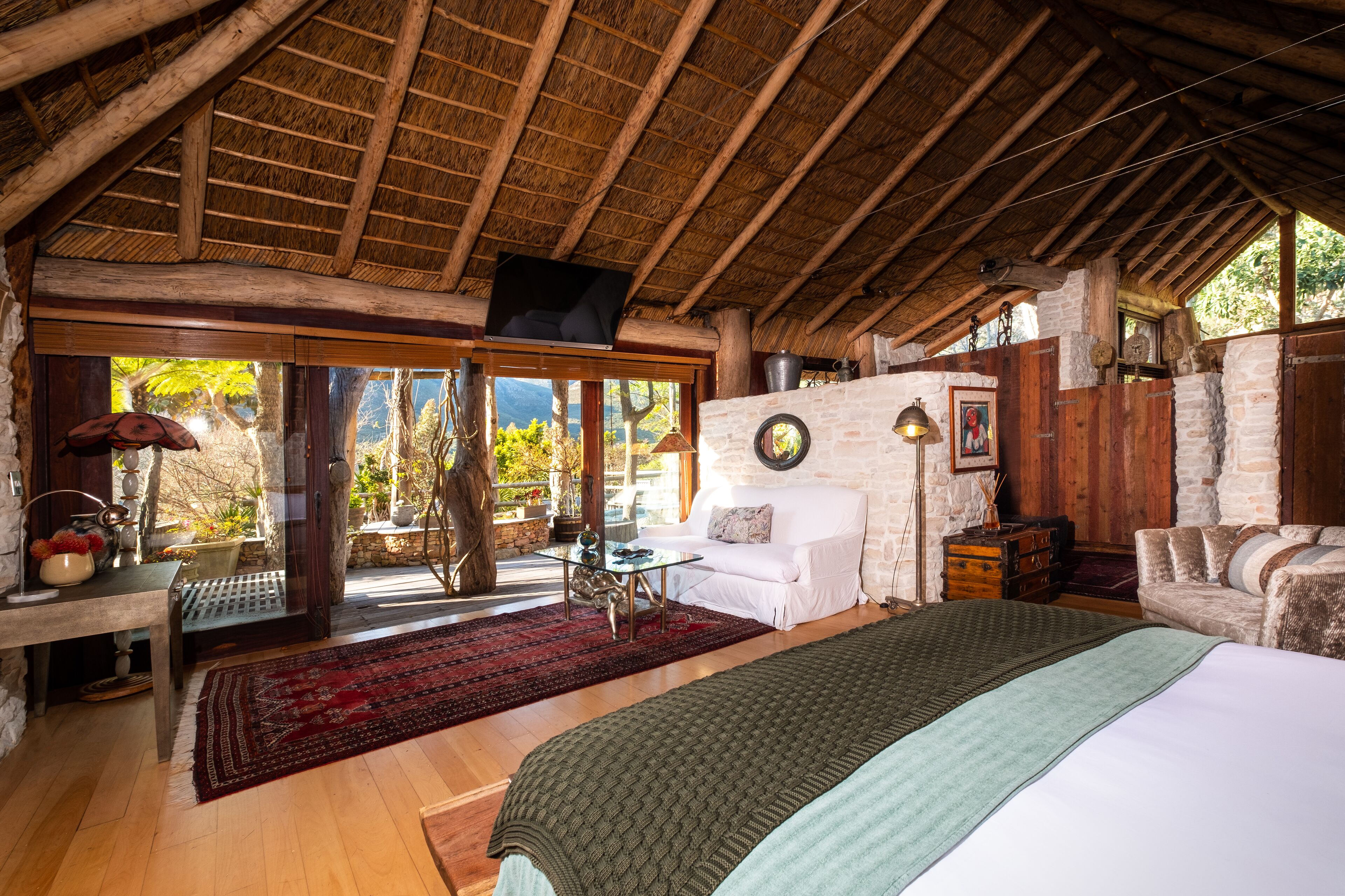 A rustic wooden room with a bed, a sofa, a dining table, and a TV mounted on the ceiling.