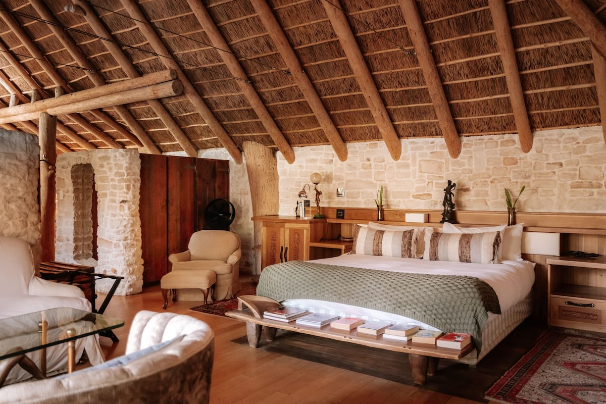 Ein gemütliches Zimmer mit Holzbalkendecke, Steinwänden, einem Bett mit grüner Decke, einem Nachttisch aus Holz und einem Sessel.