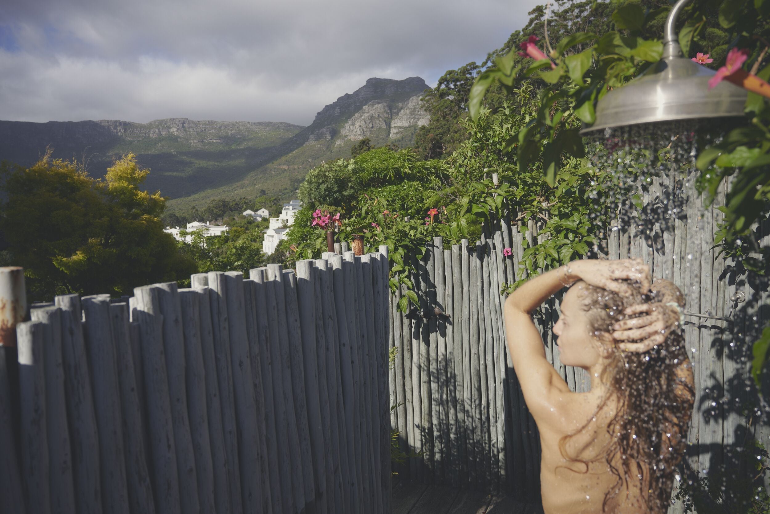 Eine Person in einem Gartenbereich mit Holzzaun, Duschkopf aus Metall und einer bergigen Landschaft im Hintergrund.