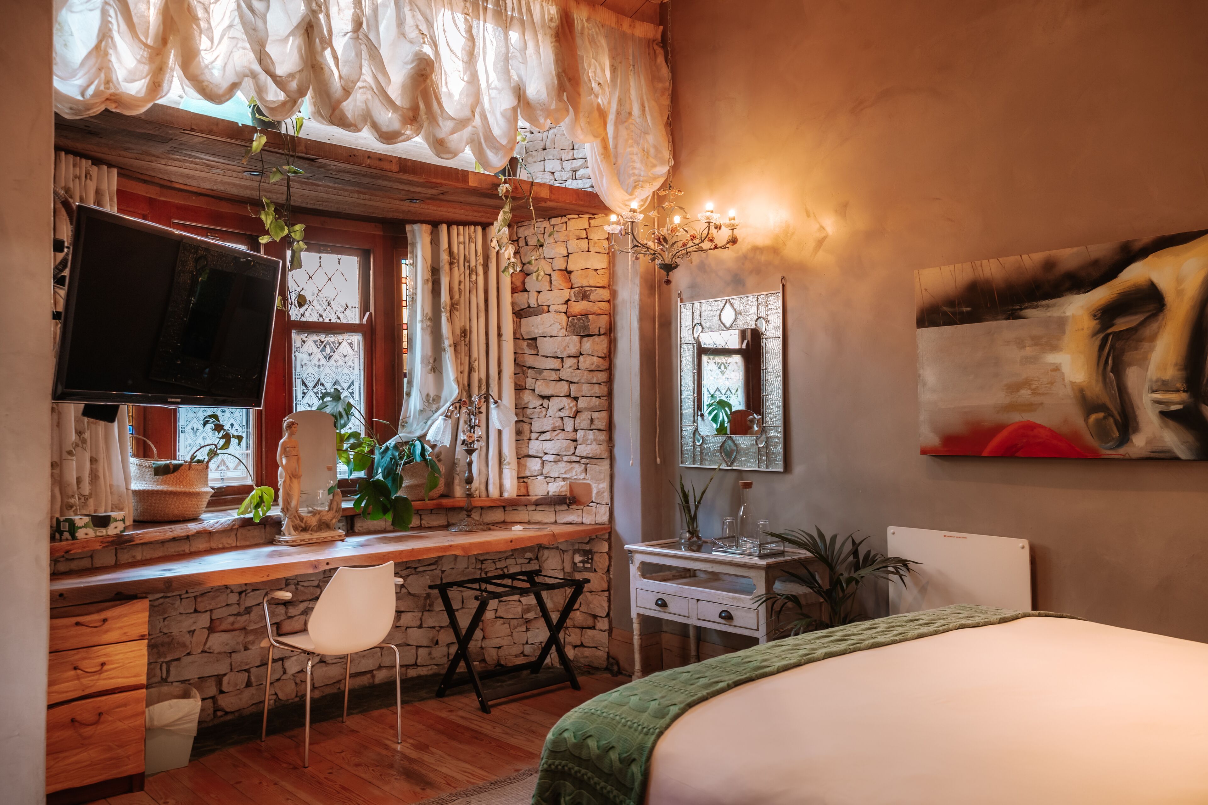 A cozy room with a stone wall, a wooden desk, a white chair, a bed, and a painting on the wall.