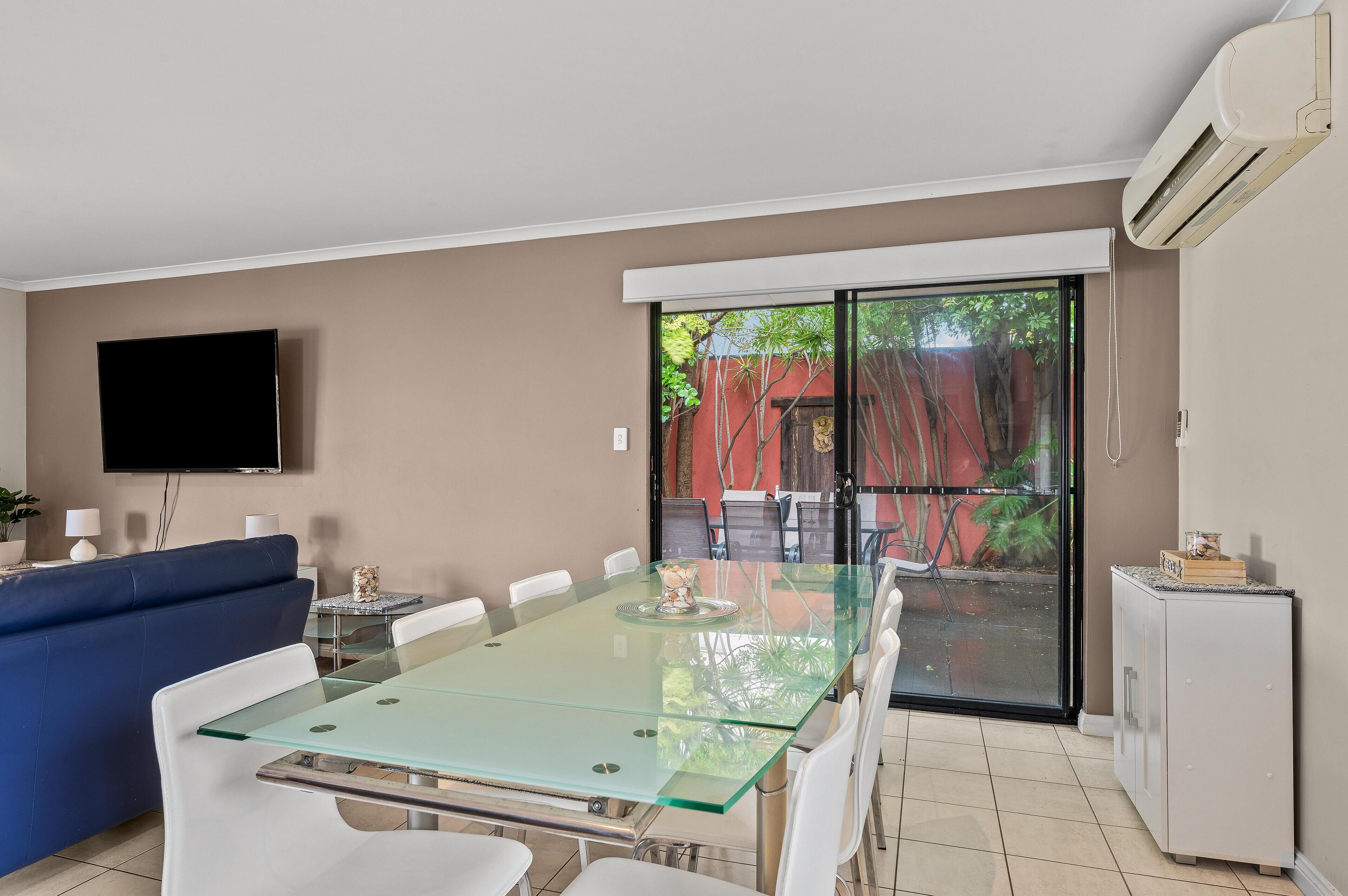 A modern dining area with a glass table, white chairs, a blue sofa, and a TV mounted on the wall.