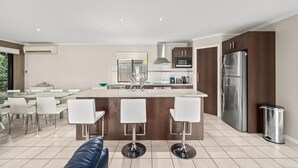 A modern kitchen with a central island, stainless steel appliances, and a dining area with white chairs. - Seacret Oasis - Aldinga Beach - C21 SouthCoast Holidays (Aldinga Beach)