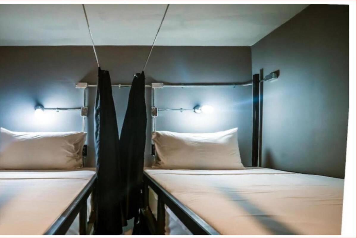 Quarto com duas camas, cada uma com travesseiro e cabeceira. Duas luminárias fixadas na parede acima de cada cama.