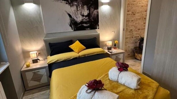 A hotel room with a bed, a nightstand, a wall-mounted artwork, and a brick wall visible through an open door.