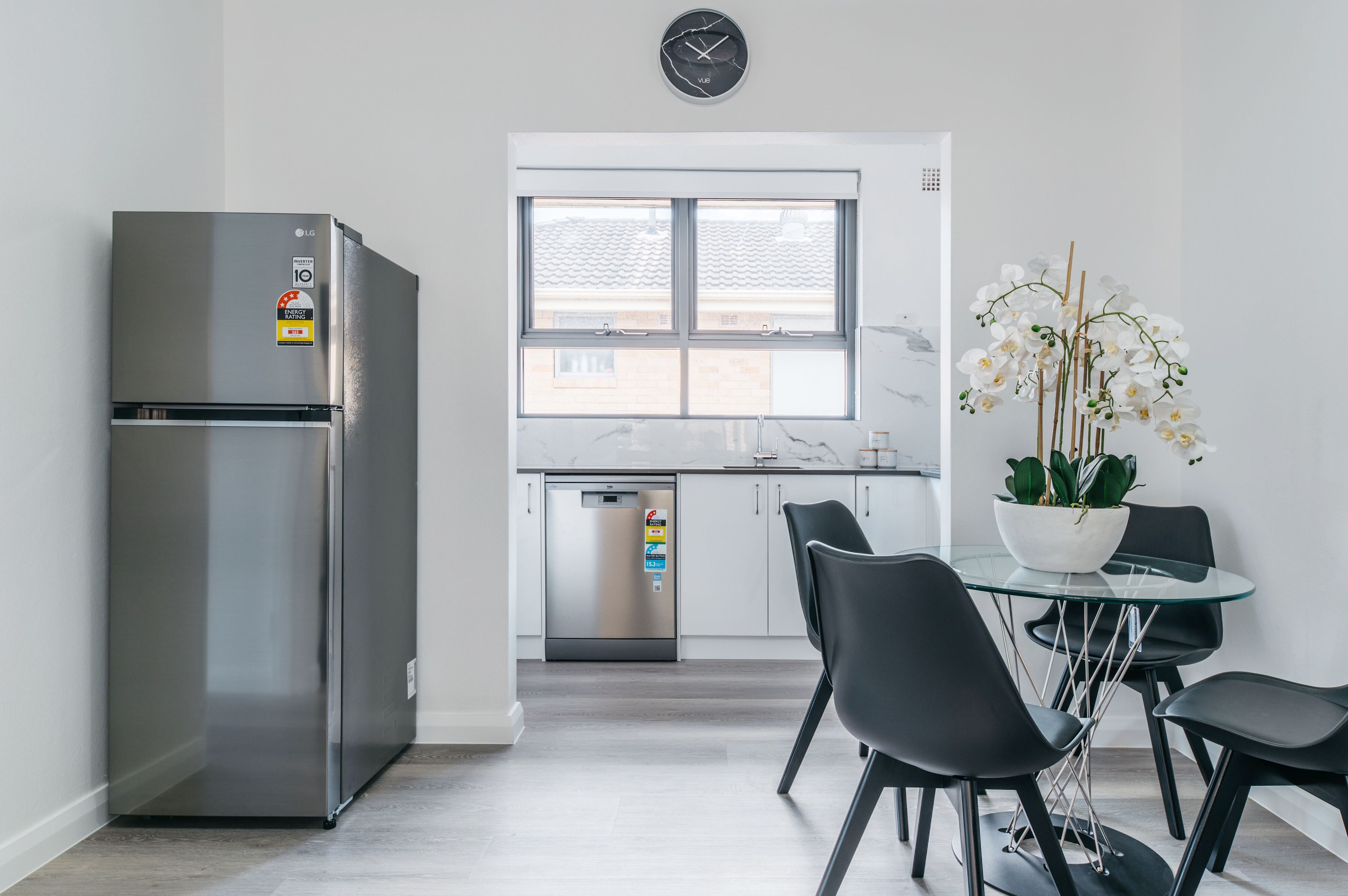 A modern kitchen with a stainless steel refrigerator, a dishwasher, and a dining area with a glass table and black chairs.
