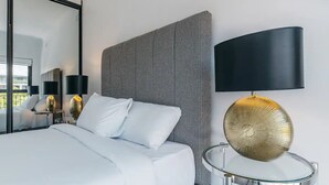 Una habitación de hotel moderna con una cama grande, un cabecero estampado, una mesita redonda de vidrio y una lámpara dorada, además de un espejo grande.