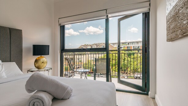 Habitación de hotel con una cama grande, una mesa redonda con sillas, un balcón con vistas a edificios y vegetación, y una lámpara fijada en la pared.