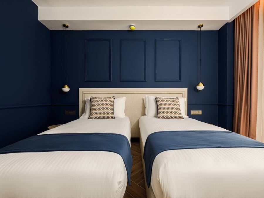 A hotel room with two beds, a white headboard, dark blue walls, and a large window with curtains.