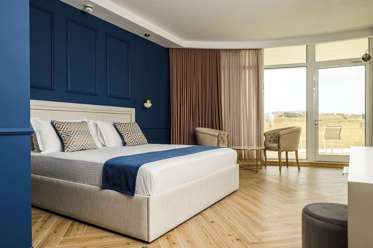A modern hotel room with a large bed, a seating area with a chair and a table, and a large window offering a view of the outdoors.