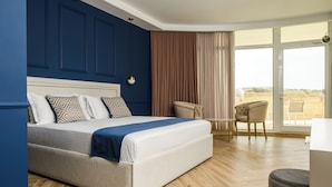 A modern hotel room with a large bed, a seating area with a chair and a table, and a large window offering a view of the outdoors.