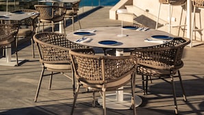 Een buiten eethoek met rieten stoelen en tafels, met uitzicht op een zwembad en de zee.