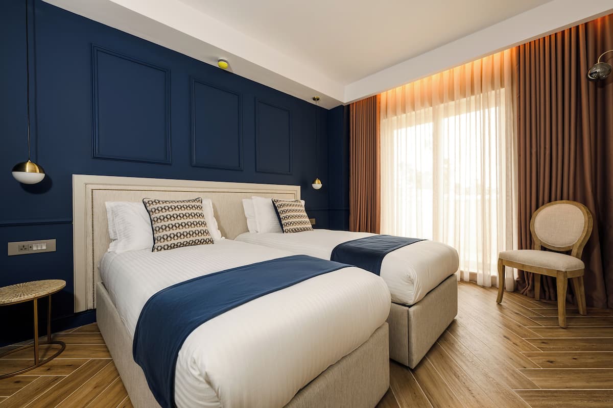 A hotel room with two beds, a small round table, a chair, and a large window with curtains.
