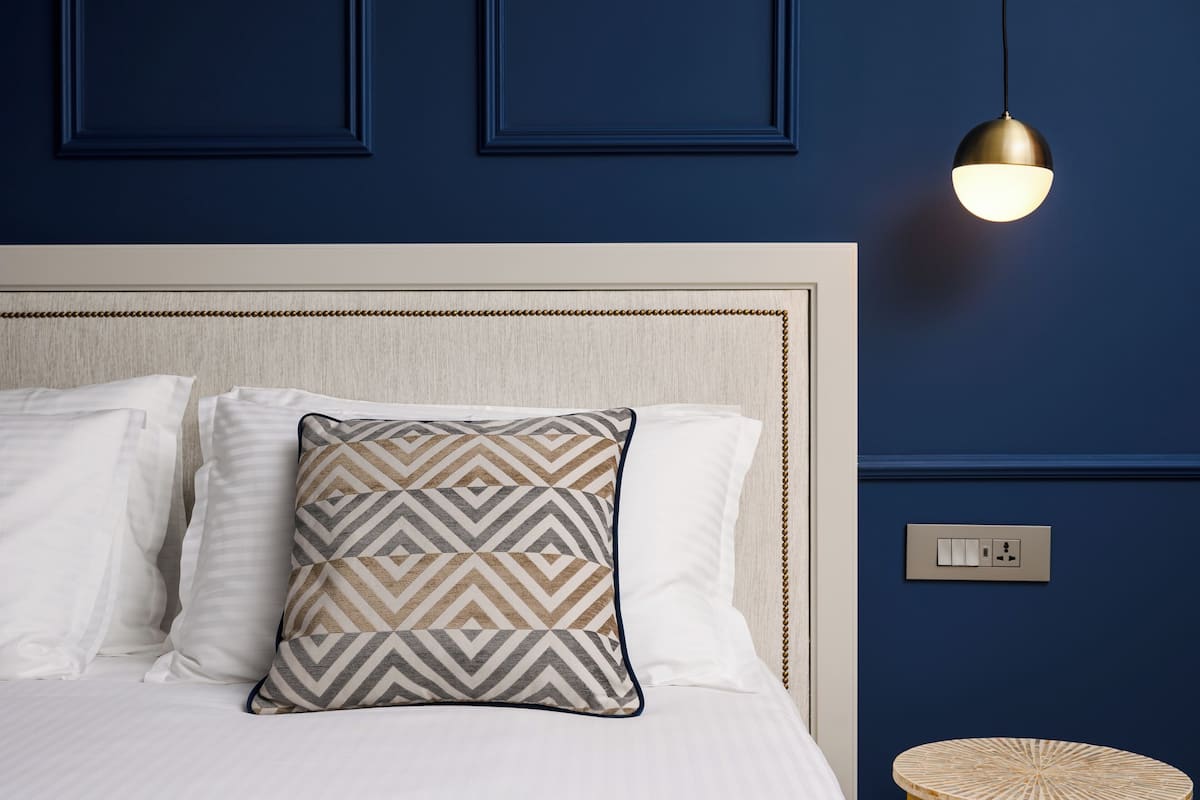 A neatly made bed with patterned pillows, a round wooden side table, and a wall-mounted light fixture.