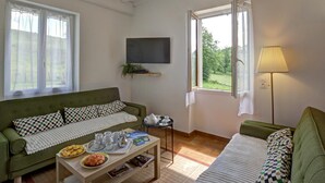 Un salon chaleureux avec un canapé vert, une table basse sur laquelle sont disposés des éléments pour le petit-déjeuner, un téléviseur fixé au mur et une fenêtre donnant sur un paysage verdoyant.