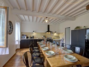 A dining area with a wooden table set for six, surrounded by chairs, with a kitchen in the background.