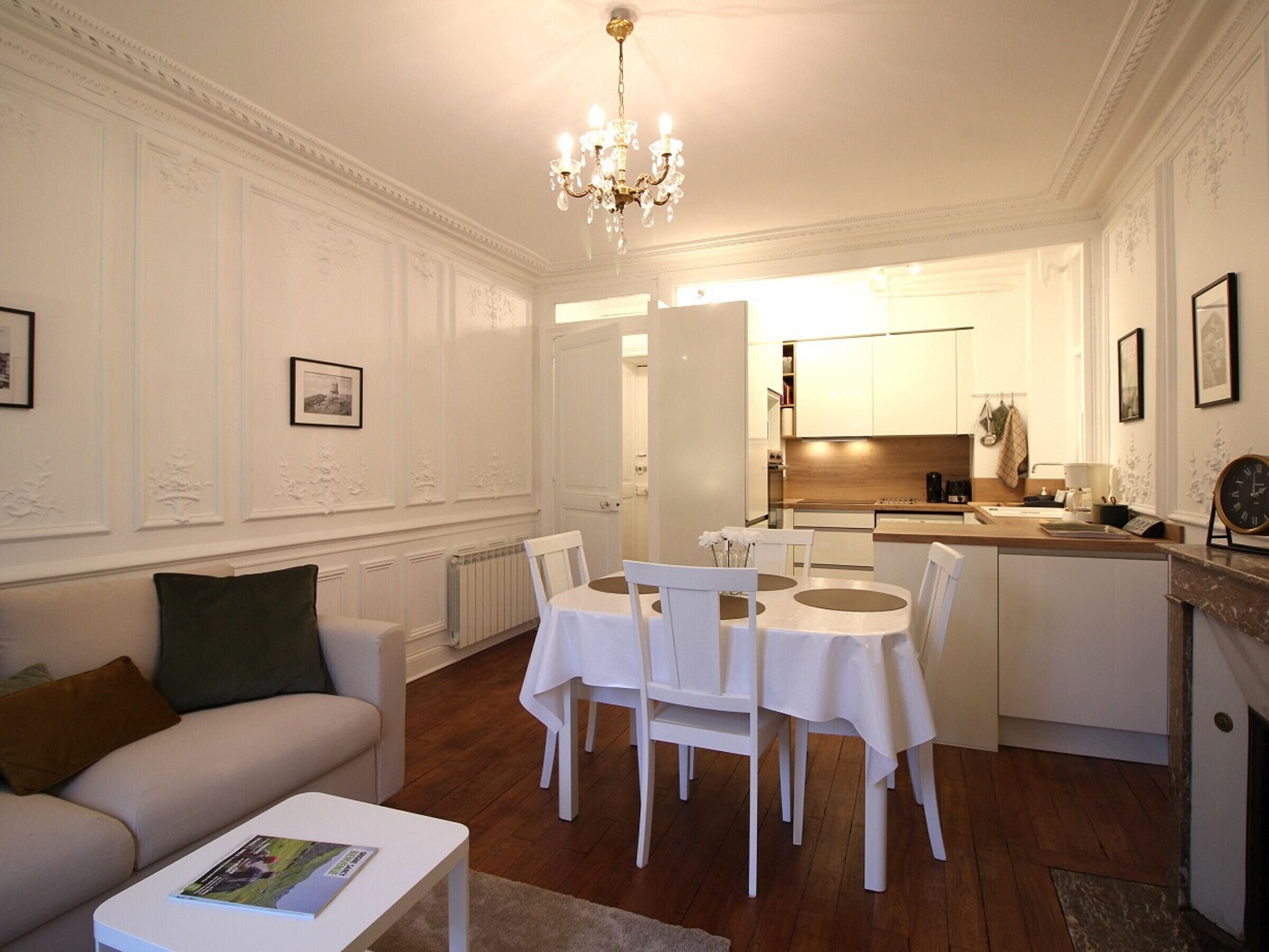 A living room with a dining table, chairs, a sofa, and a coffee table. There is a kitchen in the background with a sink, stove, and cabinets.