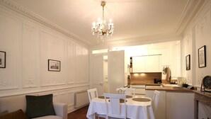 Un salon comprenant une table à manger, des chaises, un canapé et une table basse. Au fond, on aperçoit une cuisine avec un évier, une cuisinière et des placards.