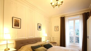 Une chambre d’hôtel avec un grand lit, deux tables de chevet avec des lampes, un lustre et une fenêtre avec des rideaux.