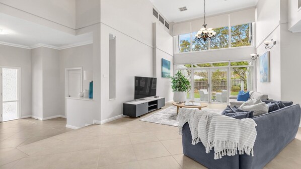 A spacious living room with a grey sofa, a TV, and a large window overlooking a garden.