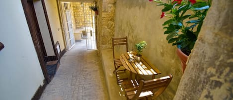 A narrow stone corridor with a wooden bench and table, a door leading to an outdoor area, and potted plants.