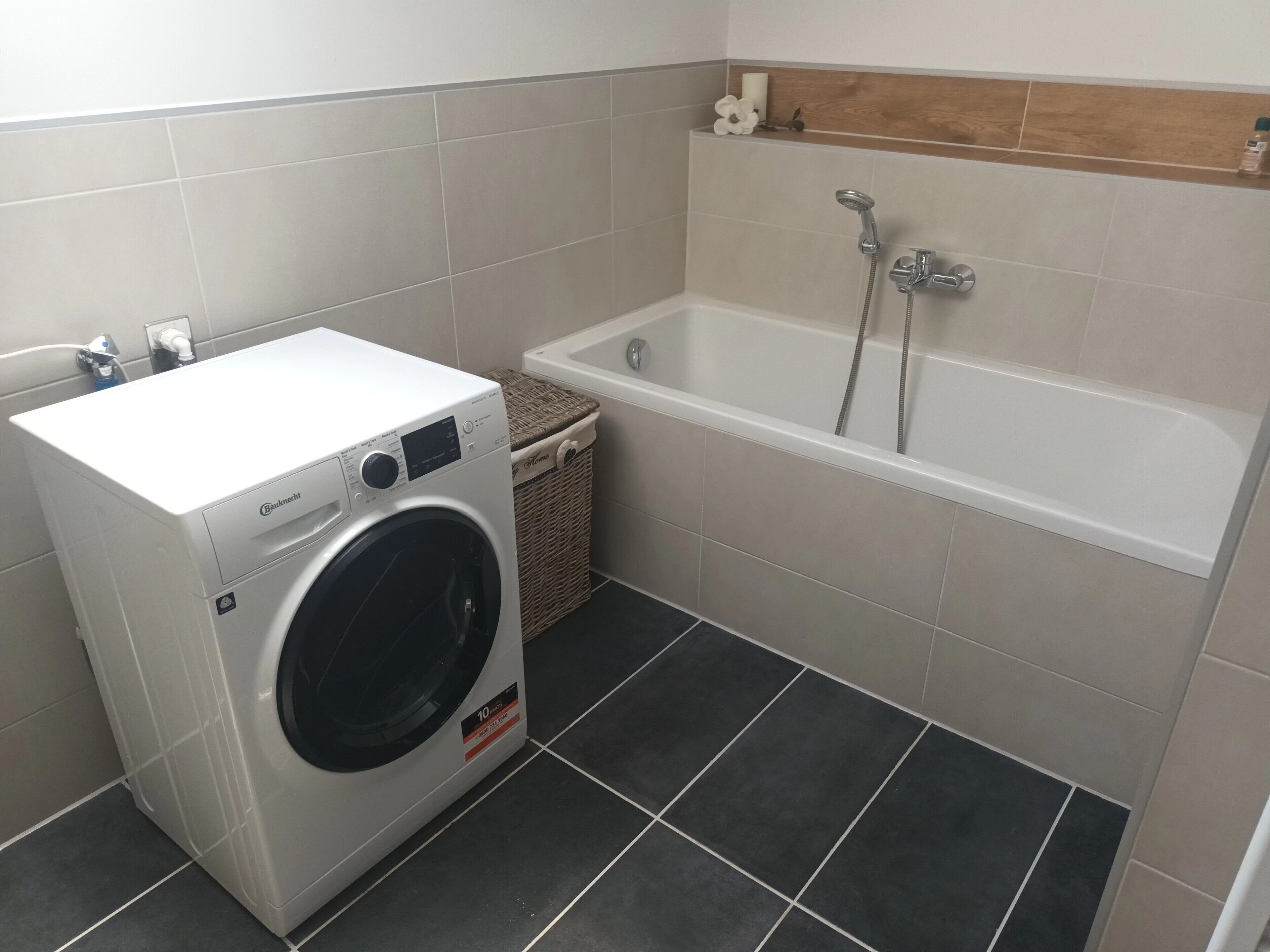 Ein Waschbereich mit Waschmaschine, Wäschekorb und Badewanne mit Duschkopf.