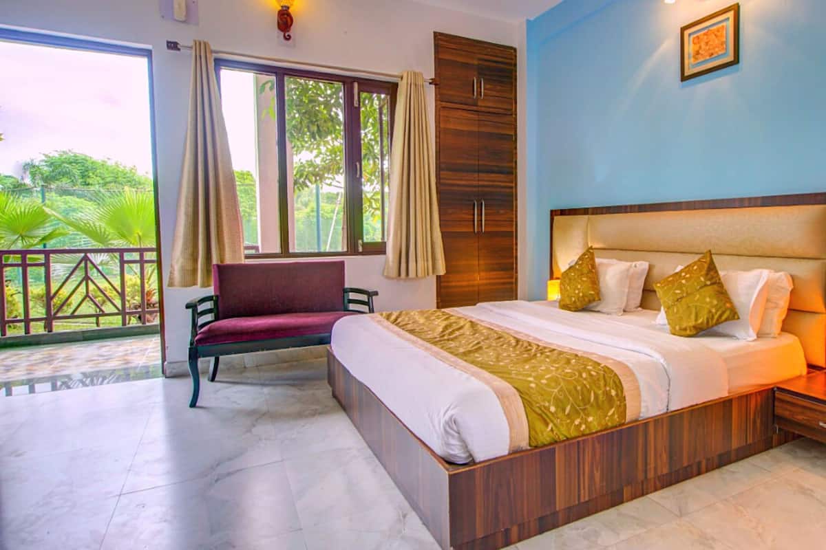 A hotel room with a large bed, a nightstand with a lamp, a chair, a wardrobe, and a balcony with a view of greenery.