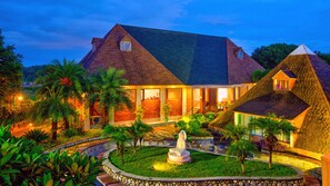 A large house with a prominent pyramid-shaped structure, a well-maintained garden with a statue, and a clear view of the surrounding landscape. - ShriGo Resort Pyramid Home Divine  (Vikasnagar)