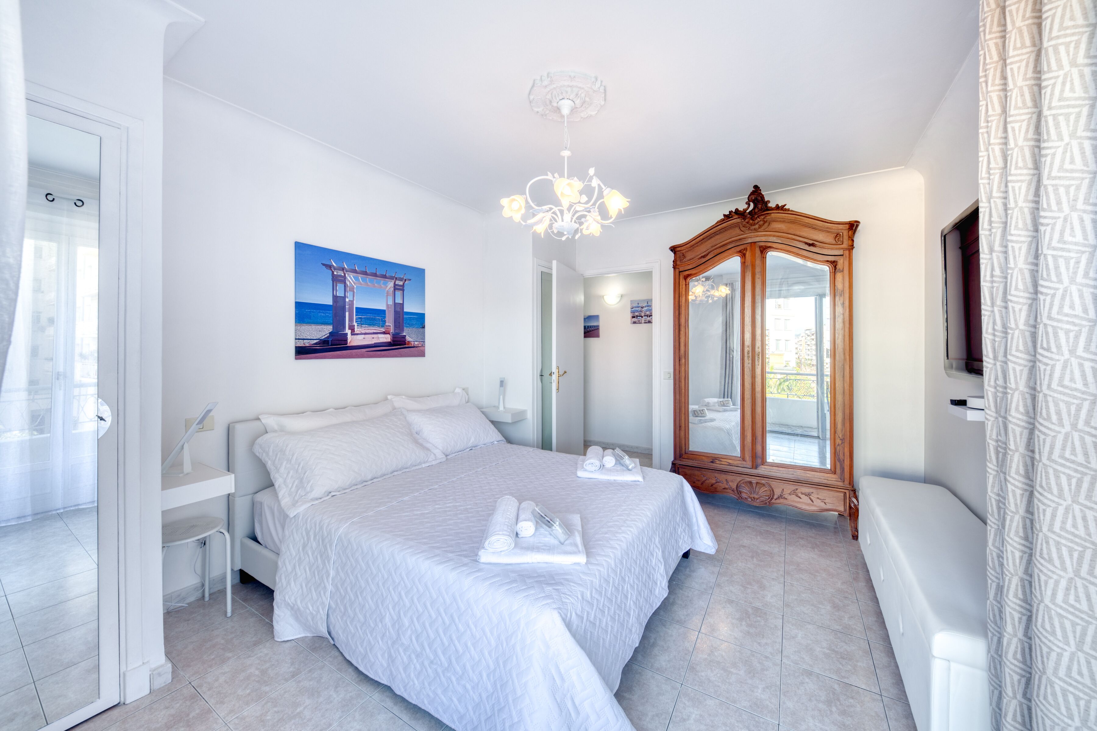 A bedroom with a bed, a wooden wardrobe, a mirror, and a painting of a beach scene.