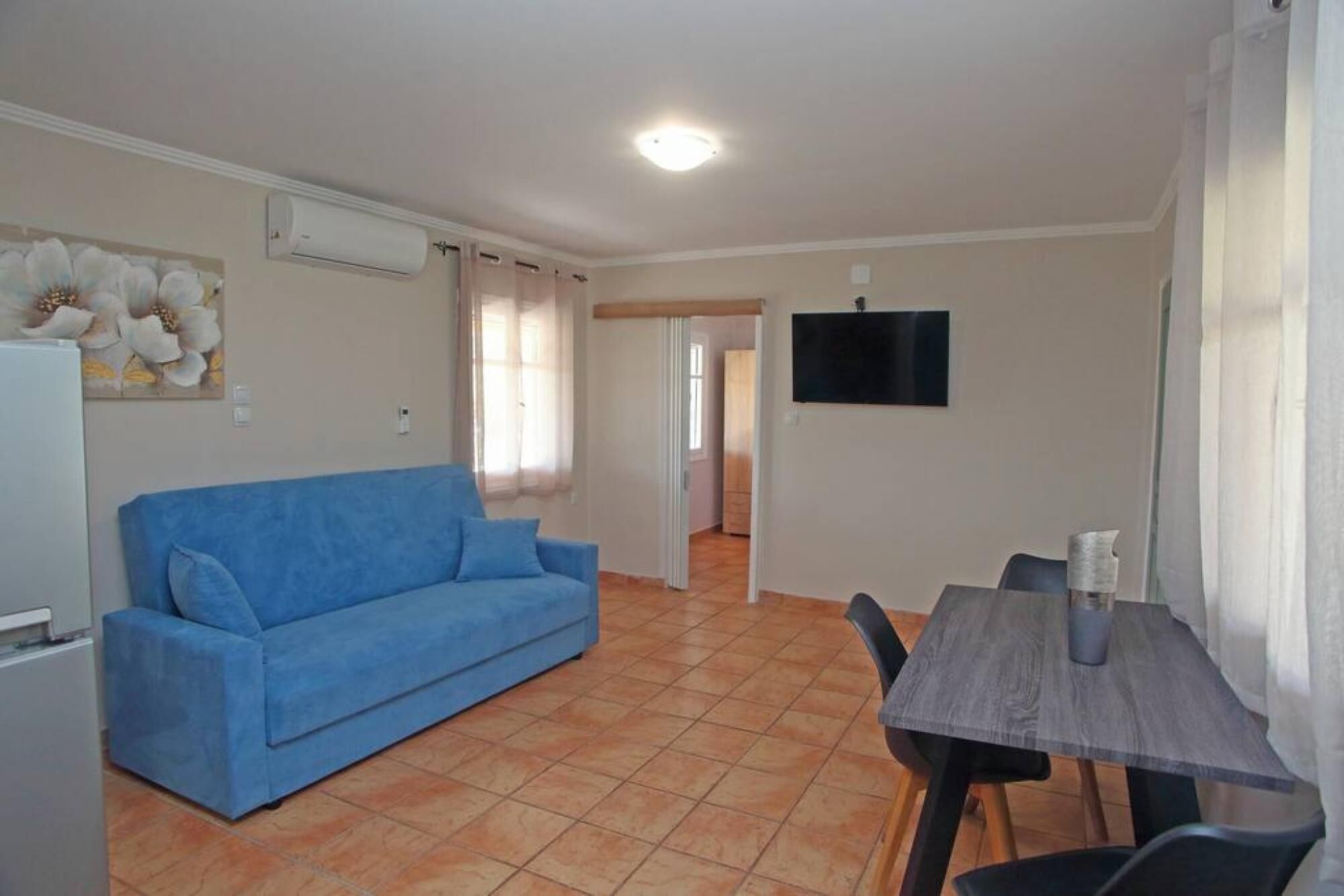 A living room with a blue sofa, a dining table with chairs, a flat-screen TV mounted on the wall, and a tiled floor.