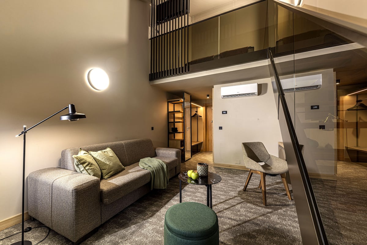 Een moderne woonkamer met een bank, een salontafel en een fauteuil. Er is een trap die naar een hoger gelegen verdieping leidt.