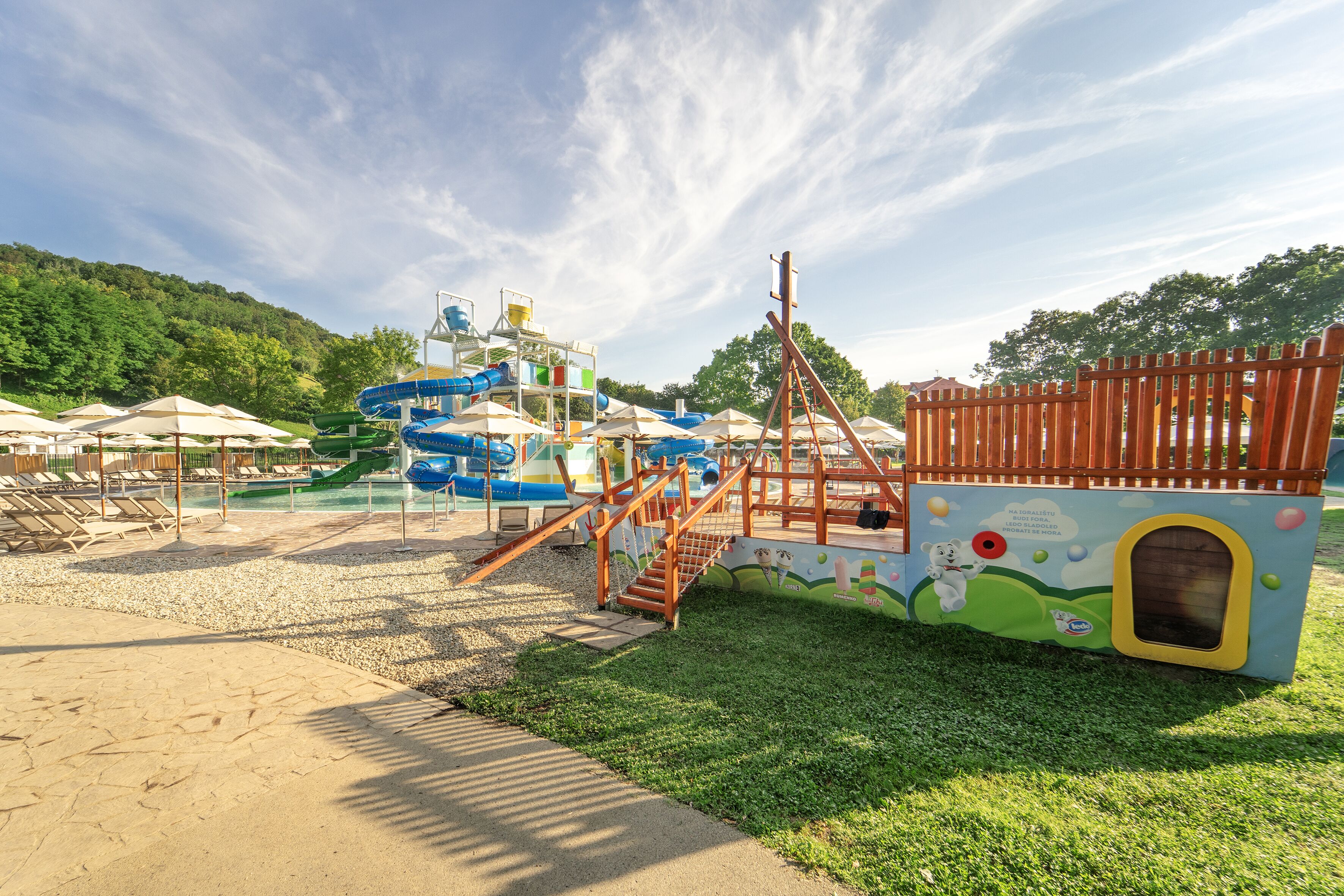 A playground with slides and climbing structures, surrounded by umbrellas and a sandy area.