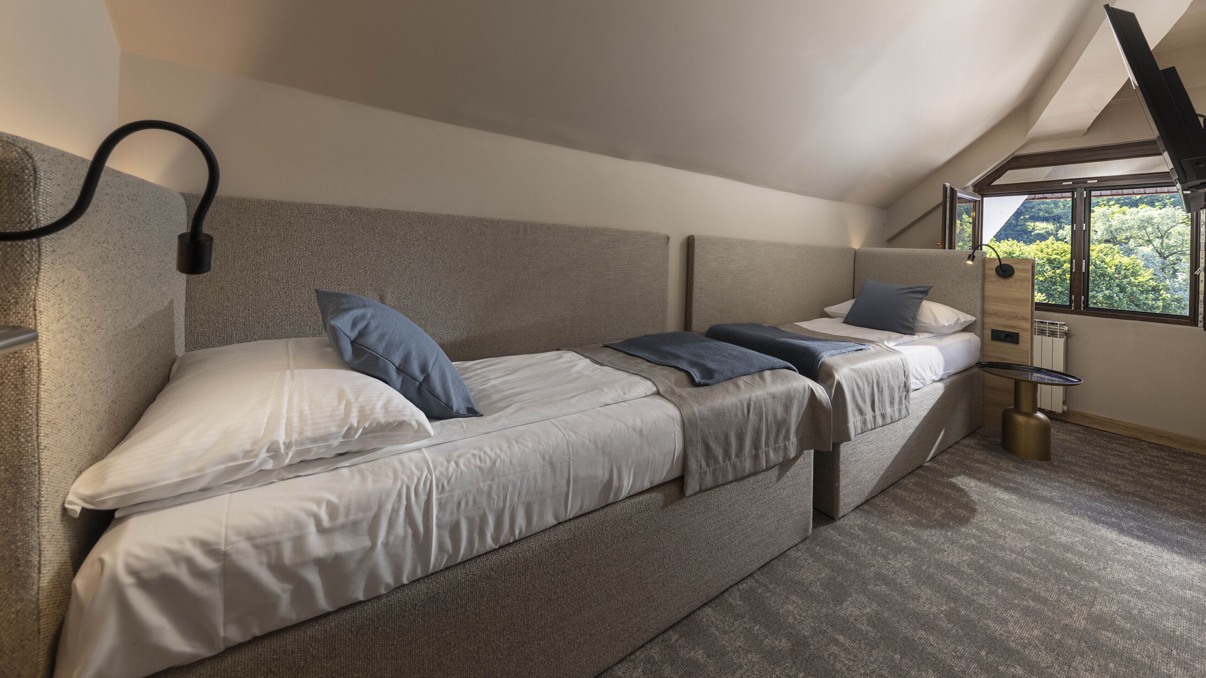 A modern bedroom with two beds, a built-in heater, and a window with a view of trees.