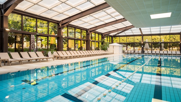 Una piscina interior con agua cristalina y azul, rodeada de sillones de descanso y amplias ventanas.