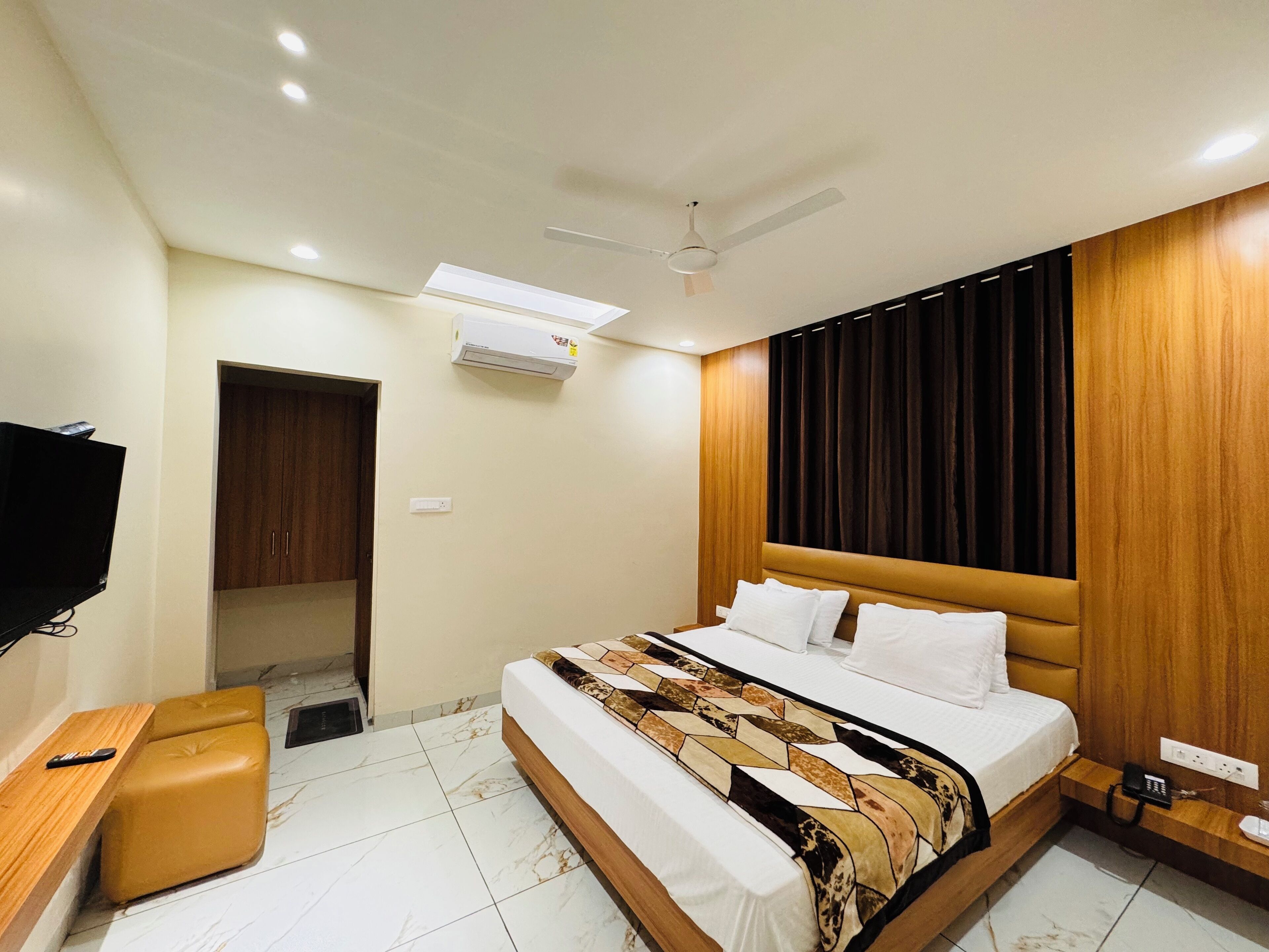 A modern hotel room with a large bed, a wooden headboard, a flat-screen TV, and a small wooden bench.