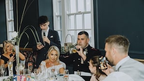 Evento formal com convidados sentados à mesa, taças de vinho, bolo e um suporte para microfone.