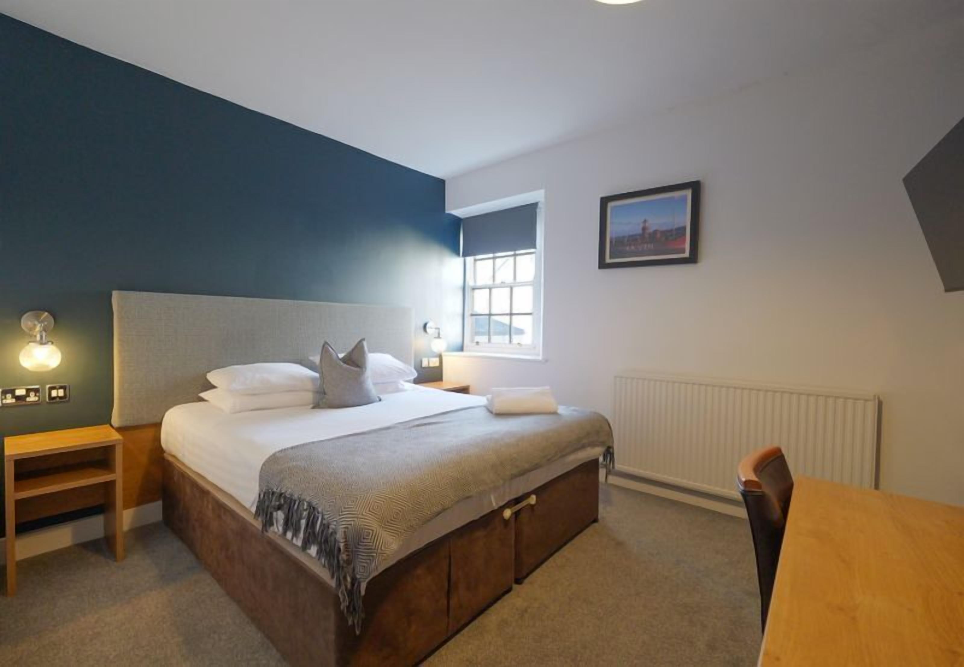 A bedroom with a bed, bedside table, lamp, window, and a framed picture on the wall.