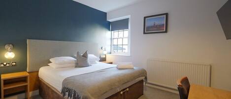 A bedroom with a bed, bedside table, lamp, window, and a framed picture on the wall.