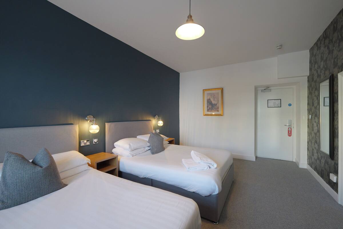 Habitación de hotel con dos camas, una mesita de noche con lámpara, un cuadro enmarcado en la pared y una puerta con extintor.