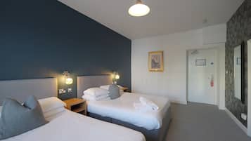 Une chambre d’hôtel avec deux lits, une table de chevet avec une lampe, un tableau encadré au mur et une porte avec un extincteur.