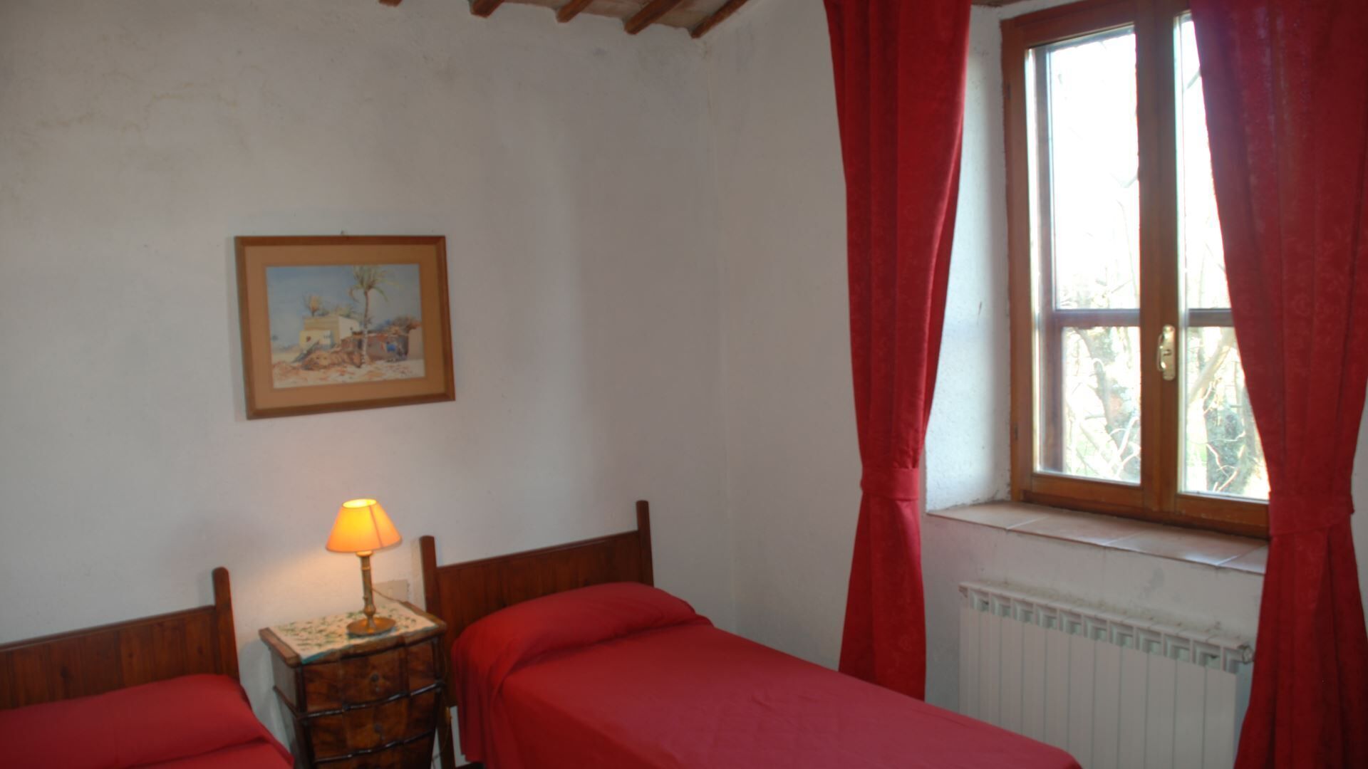 Een kamer met twee enkele bedden, een nachtkastje met lamp, een schilderij aan de muur en een raam met rode gordijnen.