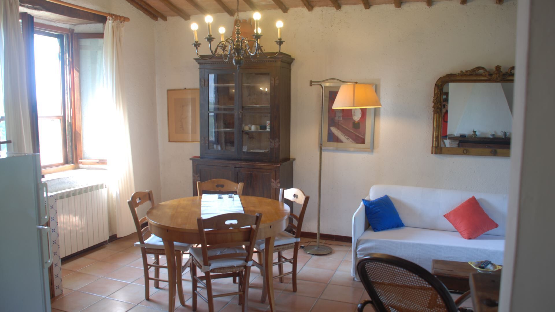 A dining area with a wooden table and chairs, a cabinet with glass doors, a floor lamp, and a sofa with cushions.
