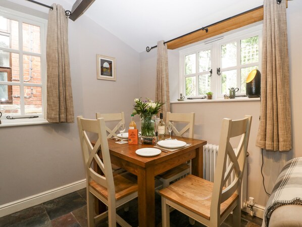 A dining area with a wooden table set for four, surrounded by wooden chairs. There is a window with curtains, a radiator, and a small table with a lamp and a potted plant. - The Old Bakehouse (Leominster)