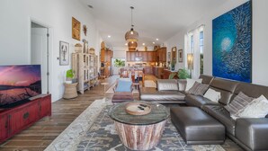 A spacious living room with a large sectional sofa, a glass coffee table, and a flat-screen TV. There is a kitchen in the background with wooden cabinets and a dining area. - Tranquil Tides, an exquisite beach home on Hill St with beach access directly across street! (New Smyrna Beach)